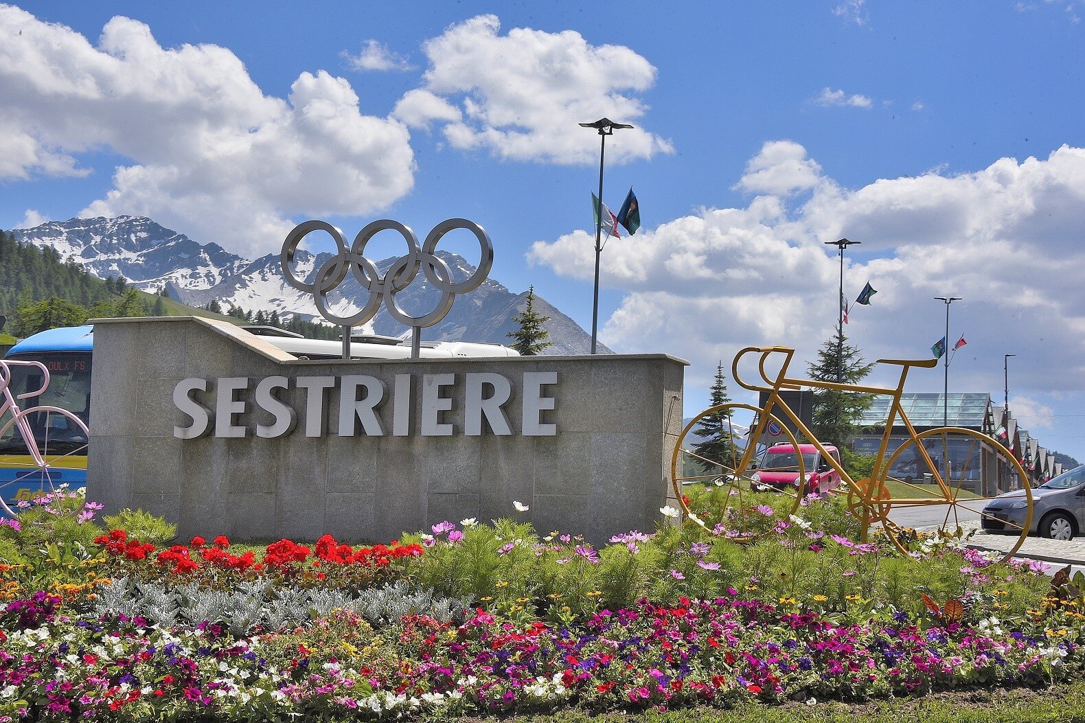 Vibrant scene with "SESTRIERE" sign, Olympic rings sculpture, flowerbed, bicycle sculpture, flags, mountains, and road.